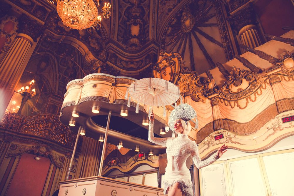 Performer in white costume poses beneath ornate baroque ceiling with grand chandeliers.