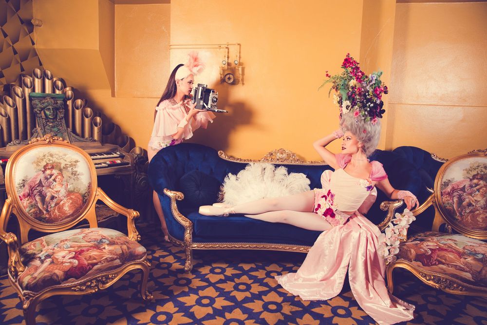 Two performers in elegant costumes lounging on vintage furniture in warm-lit theatrical setting.