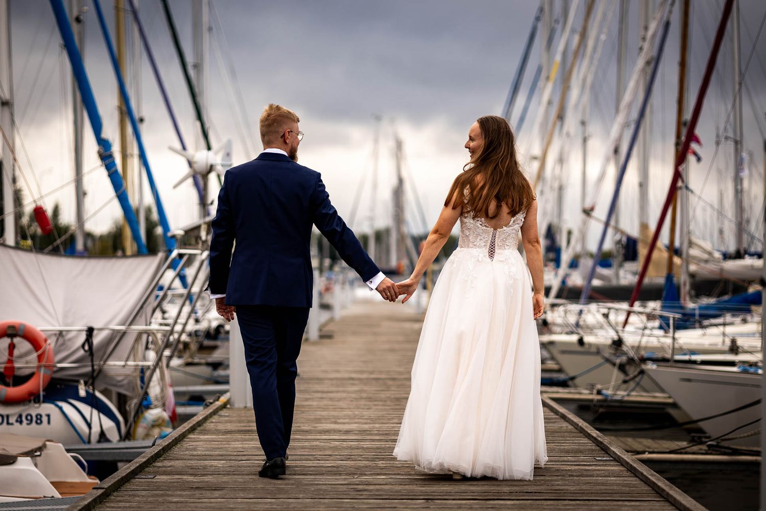 Newlyweds walking along a wooden dock with sailboats and marina in the background.