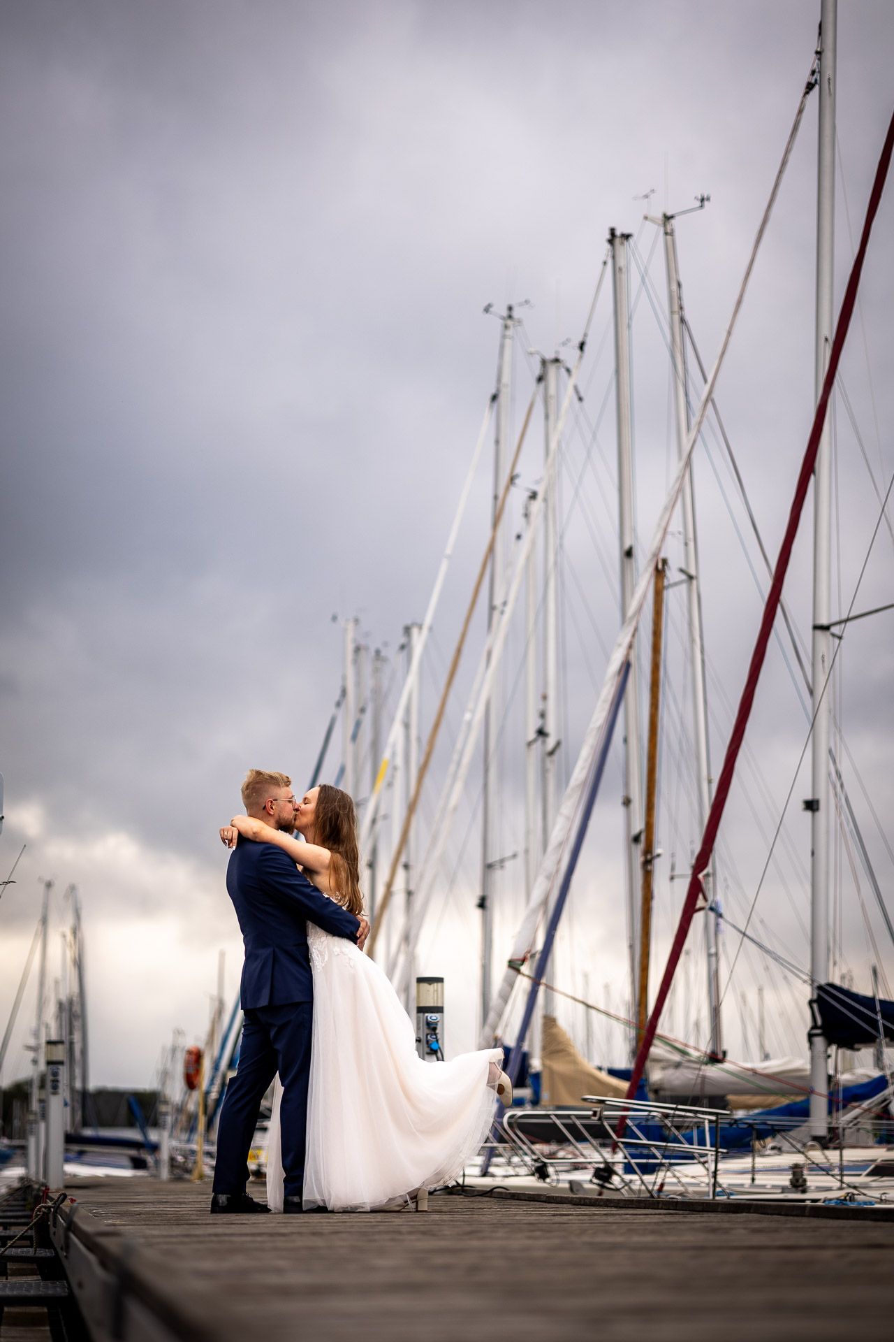 A wedding couple embraces on a wooden pier with sailboat masts towering in the background.