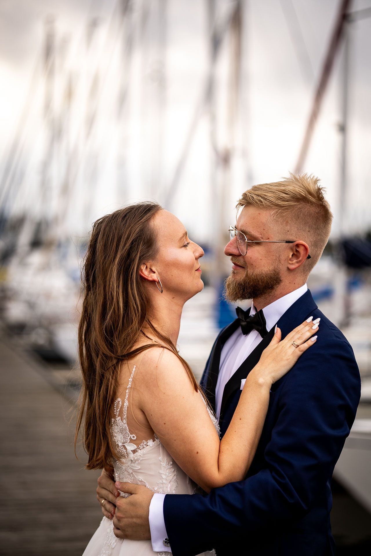 A couple shares an intimate moment on a dock surrounded by sailboats in a marina.