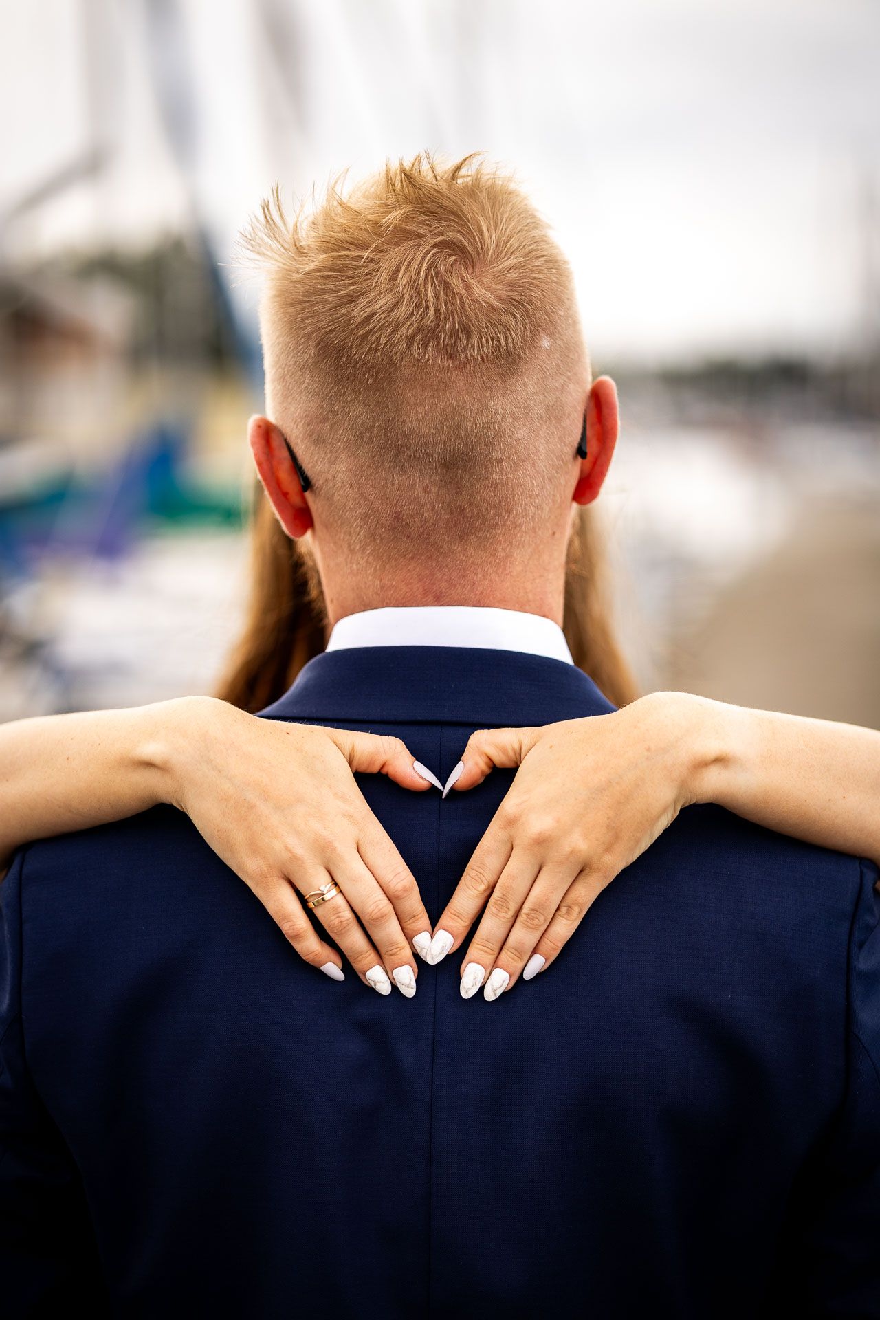 Hands with white manicured nails embrace a navy suit jacket from behind at a marina.