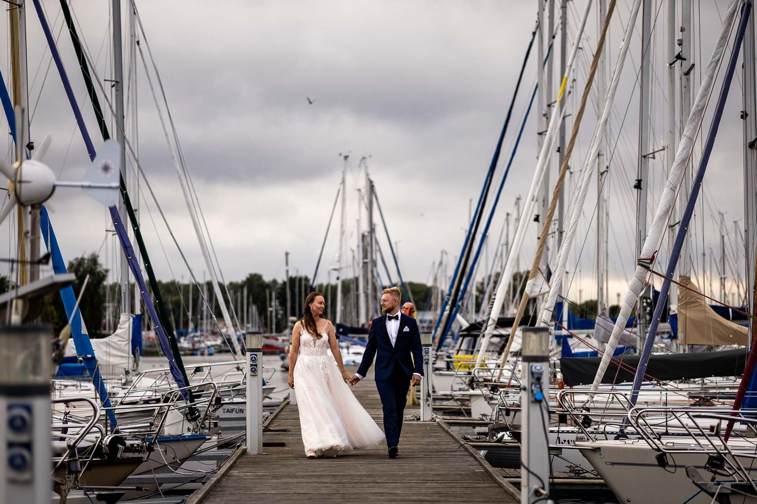 A wedding couple walks down a marina dock surrounded by moored sailboats.