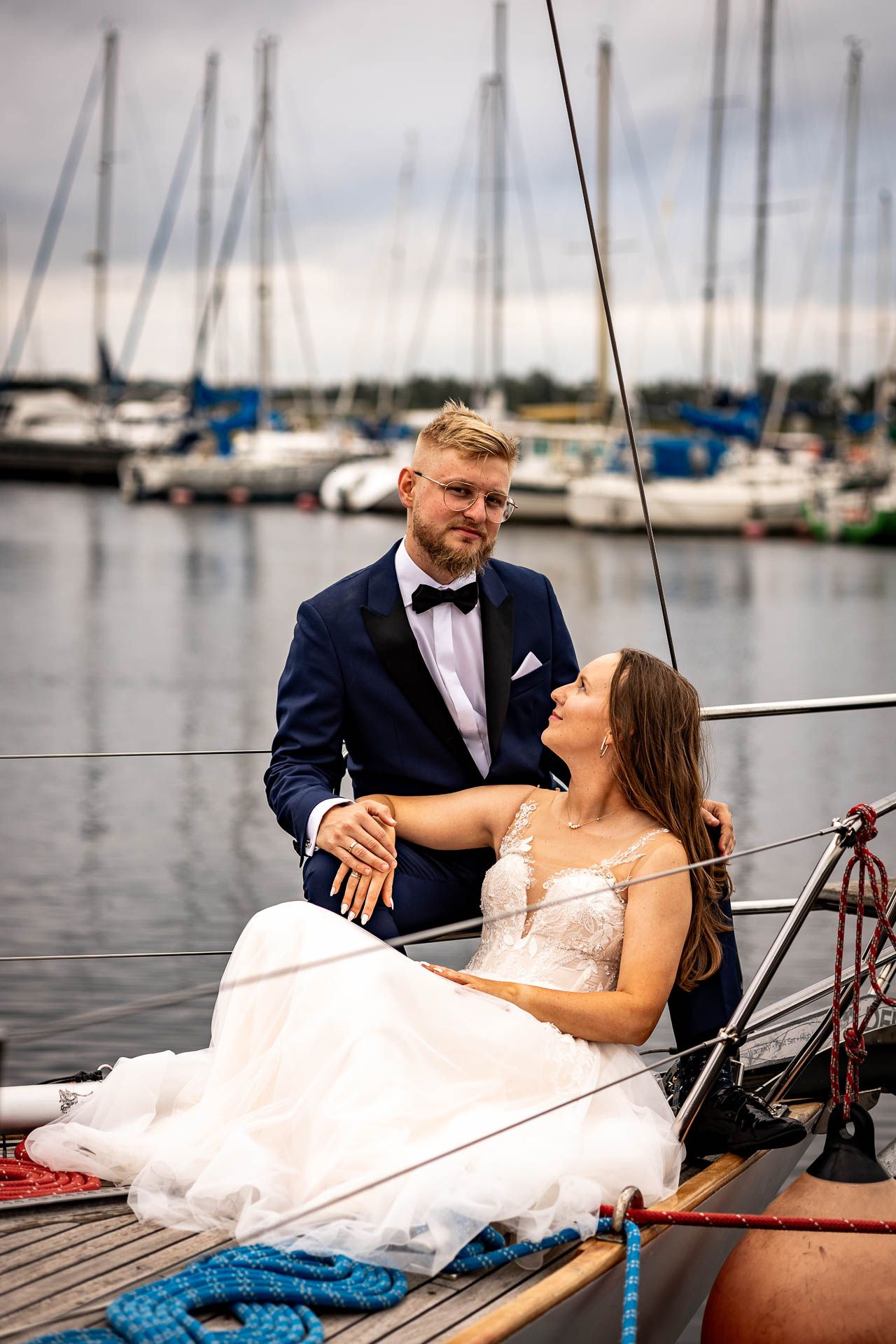 A couple relaxes on a sailboat deck at a marina, dressed in wedding attire.