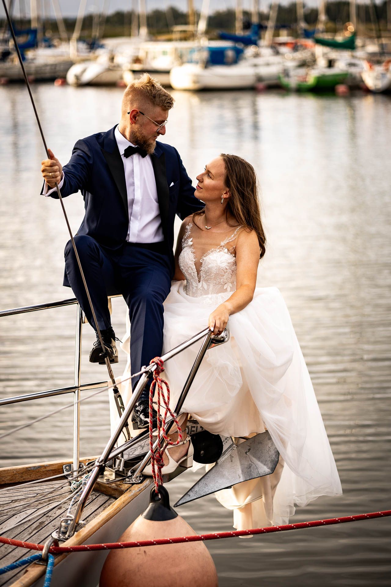 A couple in wedding attire poses on the bow of a sailboat in a marina.