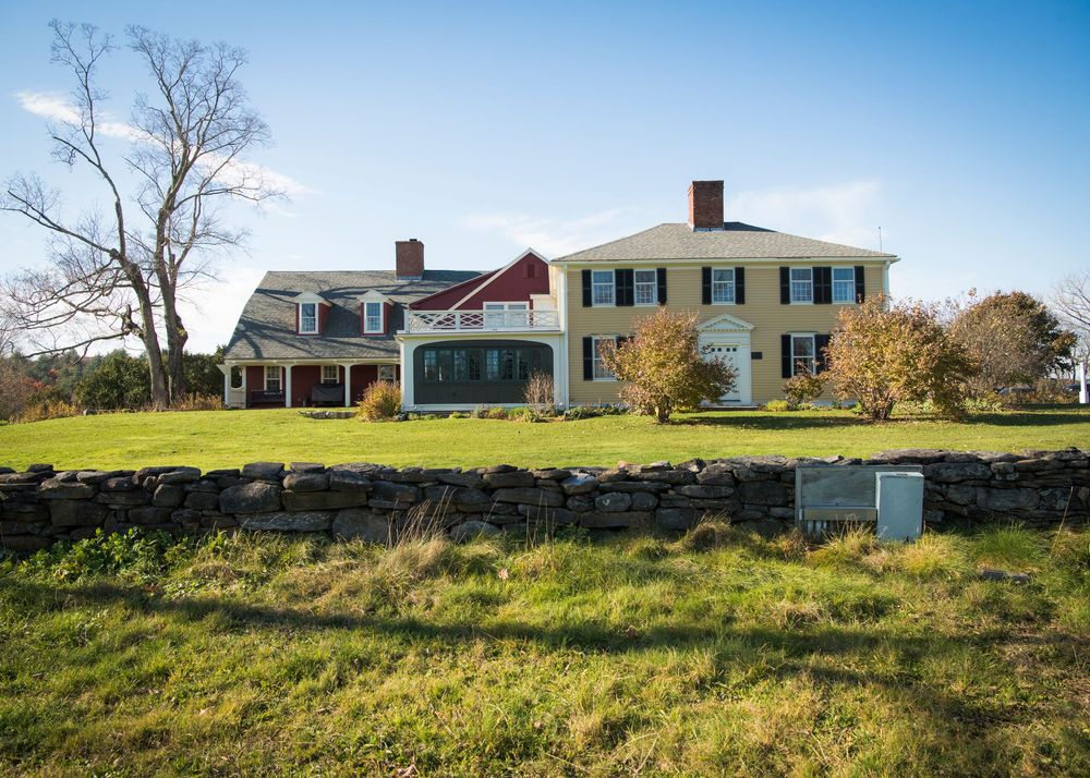 A yellow colonial style house with white trim sits on a grassy hillside under blue skies.