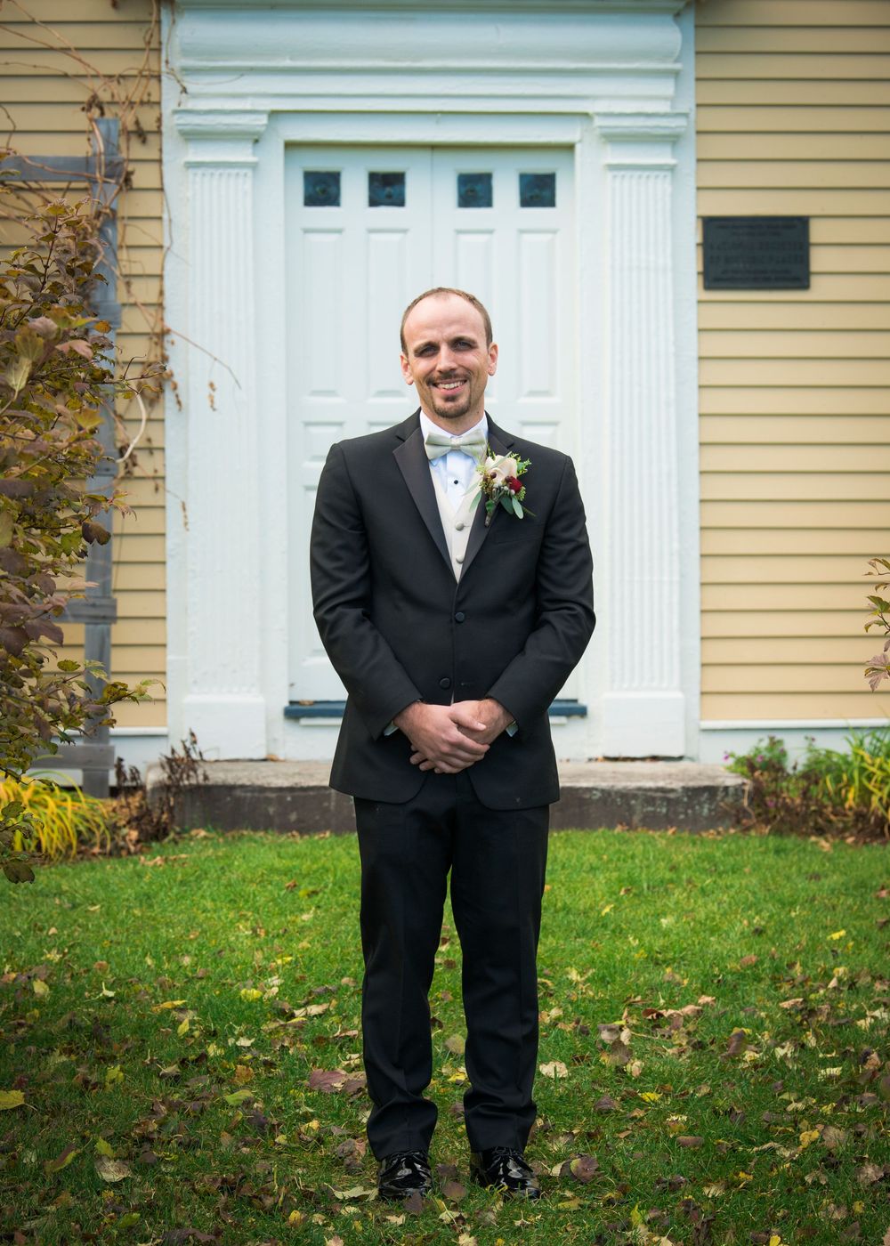Black tuxedo-clad figures stand in front of a yellow house with white doors and trim.