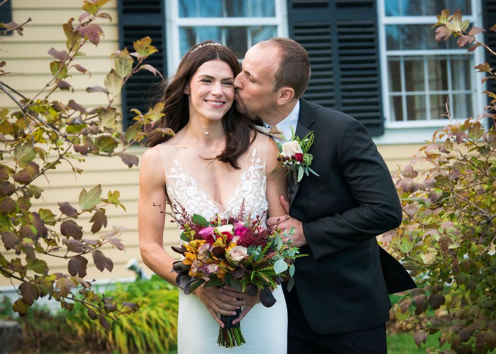 A wedding couple poses together in front of a yellow house with black shutters and autumn foliage.