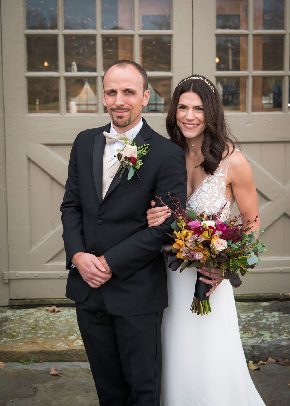 A wedding couple poses together at a rustic venue with a colorful autumn bouquet.
