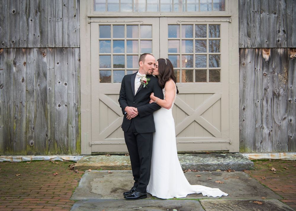 A wedding couple shares romantic moments against a rustic wooden barn wall and white door backdrop.