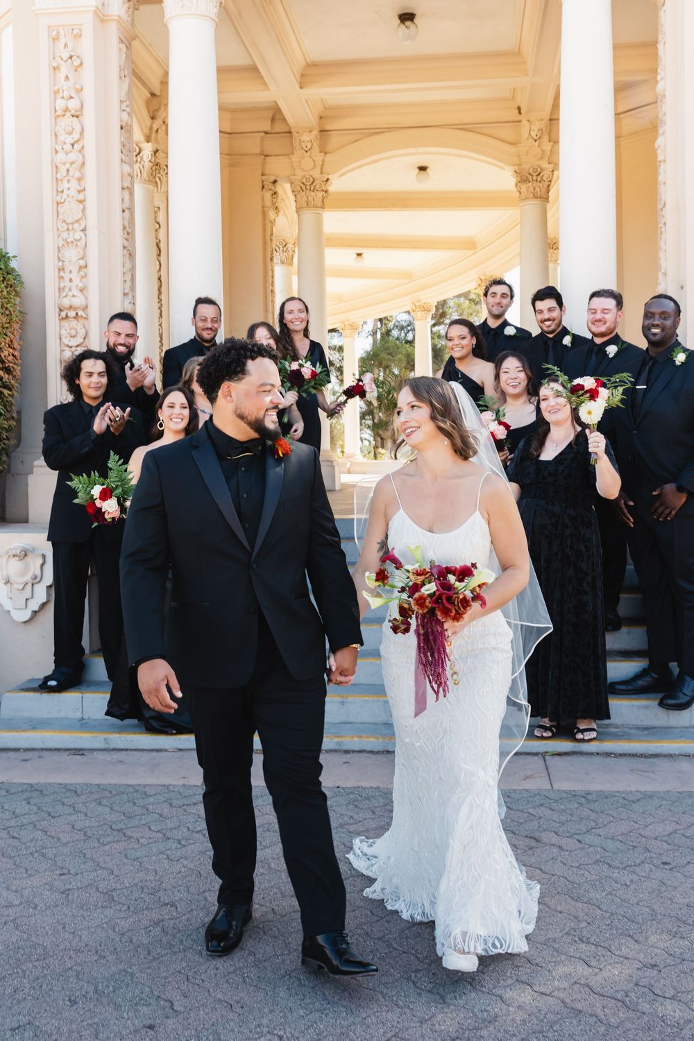 Bride and groom hold hands with wedding party in the background at Spreckels Organ Pavilion in Balboa Park