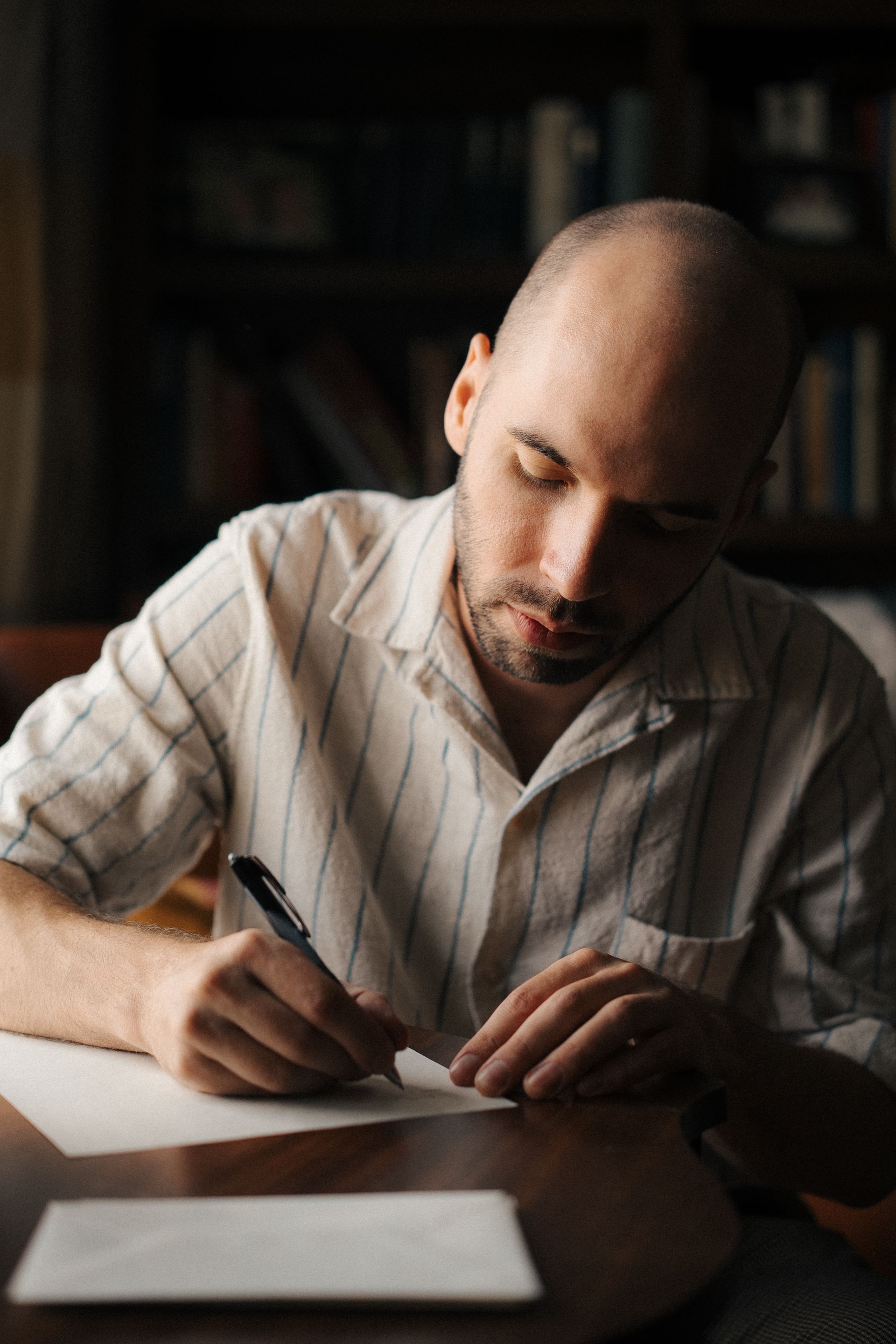 A person in a white striped shirt writes in a notebook at a desk in moody lighting.