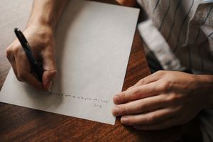 Hands writing with a fountain pen on white paper against a dark wooden desk surface.