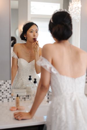 A person in a white wedding dress looks at their reflection in a bathroom mirror while getting ready.