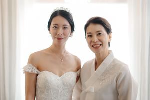 Two people pose together at a formal event, with one wearing a white wedding dress and the other in a light beige outfit.
