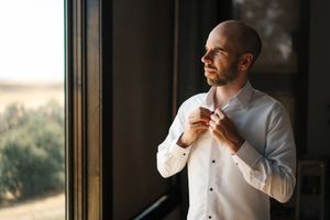 A person in a white dress shirt adjusts their collar while standing near large windows with natural light.