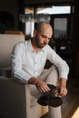 Person in white dress shirt sitting in beige chair adjusts their watch in dramatic lighting.