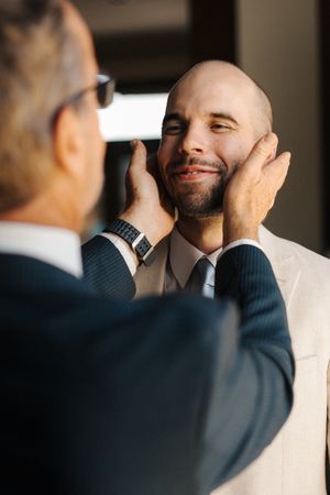 A man in a white suit gets his face touched by someone wearing a navy suit during a formal event.