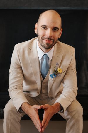 A groom in a beige three-piece suit and blue tie sits with hands clasped, wearing a yellow and blue boutonniere.