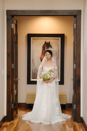 A bride in a flowing white wedding gown and veil stands elegantly in a hallway with wooden floors and neutral walls.