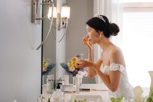 A person in a white lace dress applies makeup at a vanity with flowers and natural lighting.