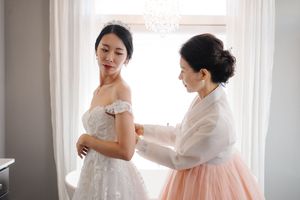 Two people prepare for a wedding ceremony as one helps adjust a beautiful white off-shoulder bridal gown by a window.