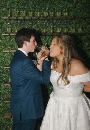 A newlywed couple shares champagne during their wedding reception against a green hedge wall.