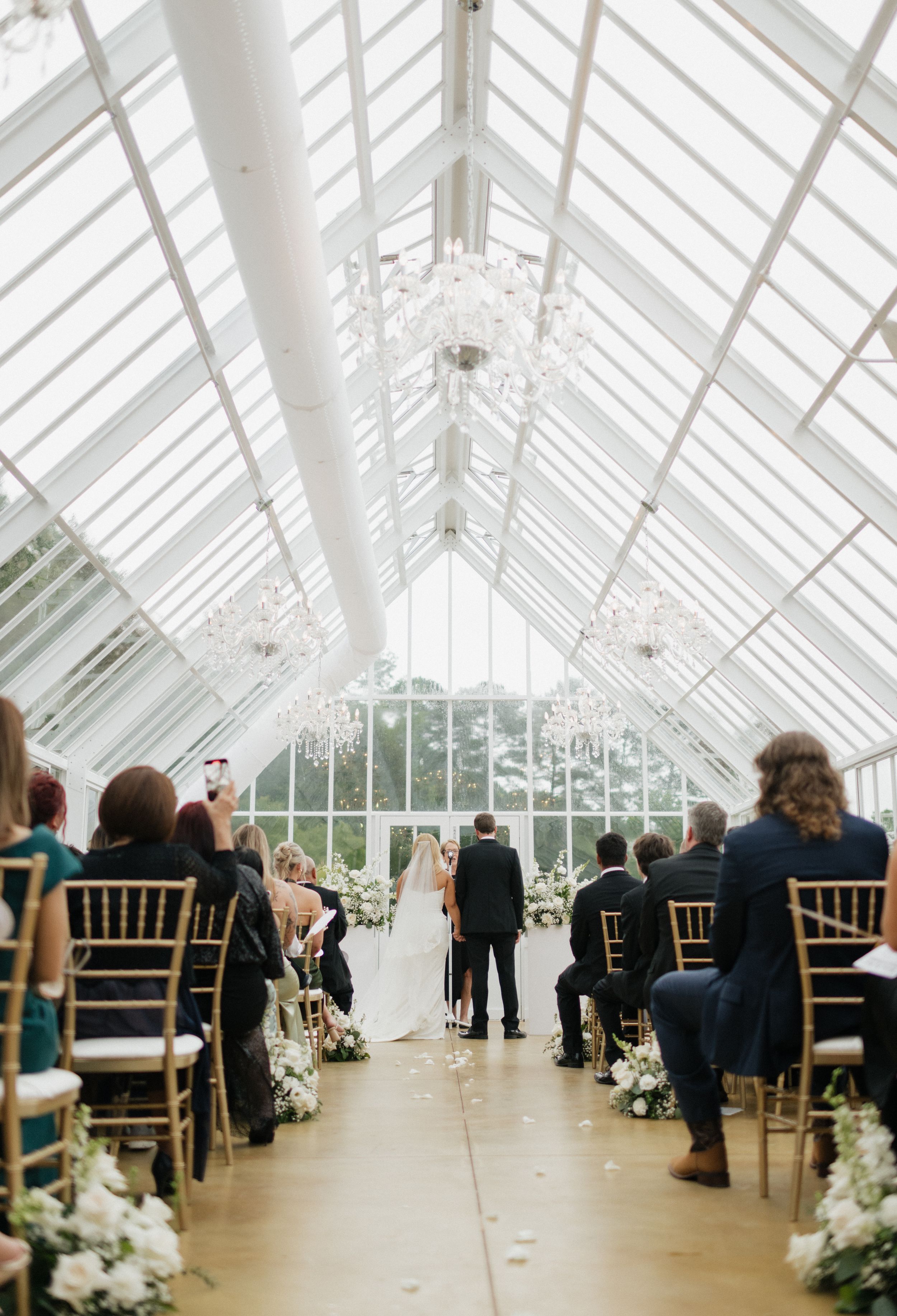 Wedding ceremony setup in an elegant conservatory with white ceiling and rows of golden chairs.