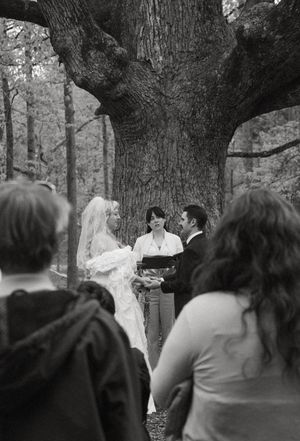 Wedding ceremony taking place in a wooded outdoor setting with guests watching.
