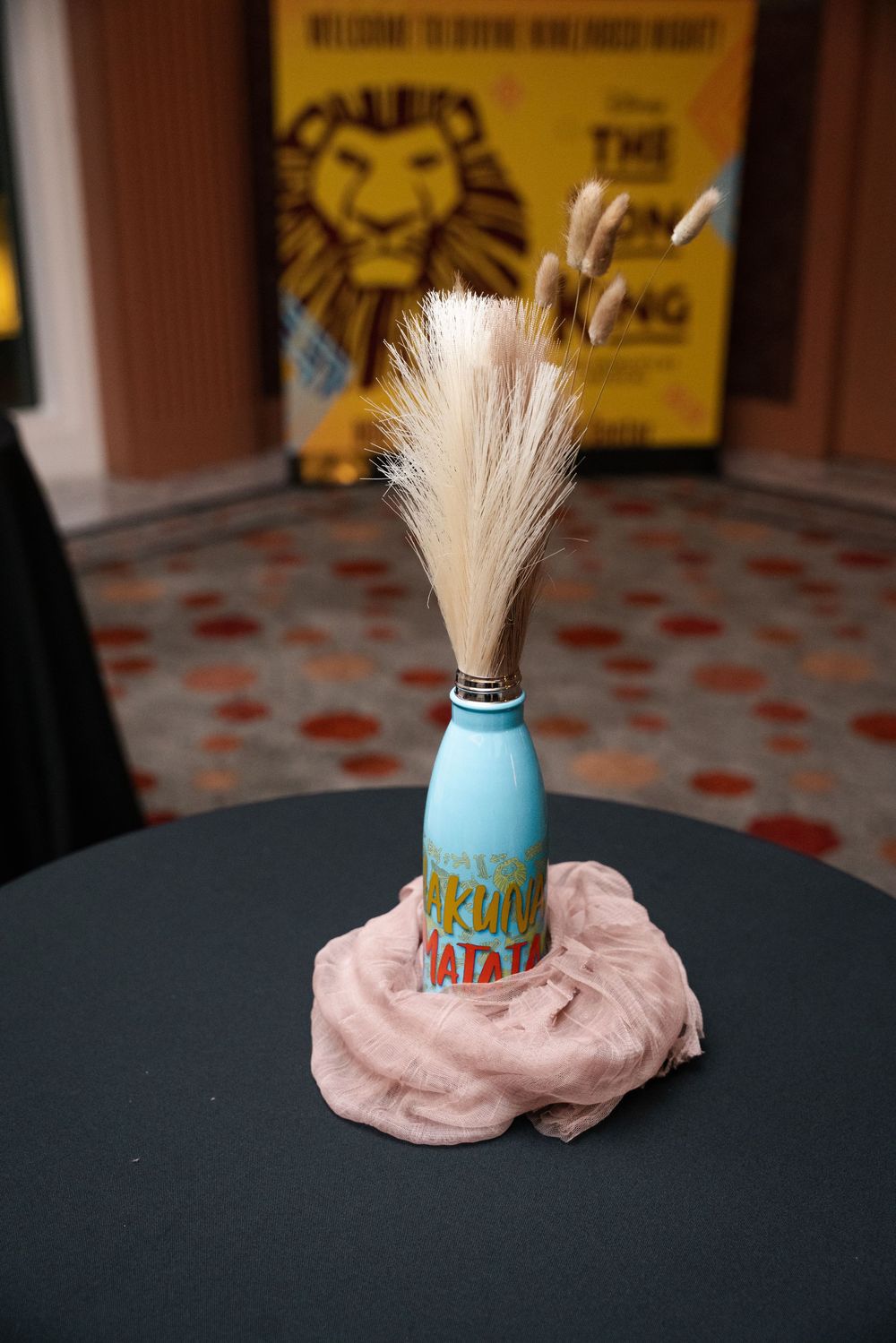 A blue glass bottle with a feathered brush top sits on a pink cloth against a dark background.