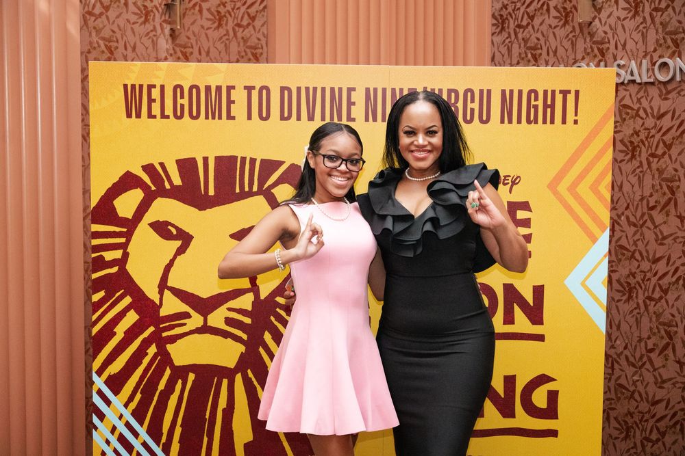 Two people pose in front of a yellow Lion King theater backdrop with Welcome text and lion logo design.