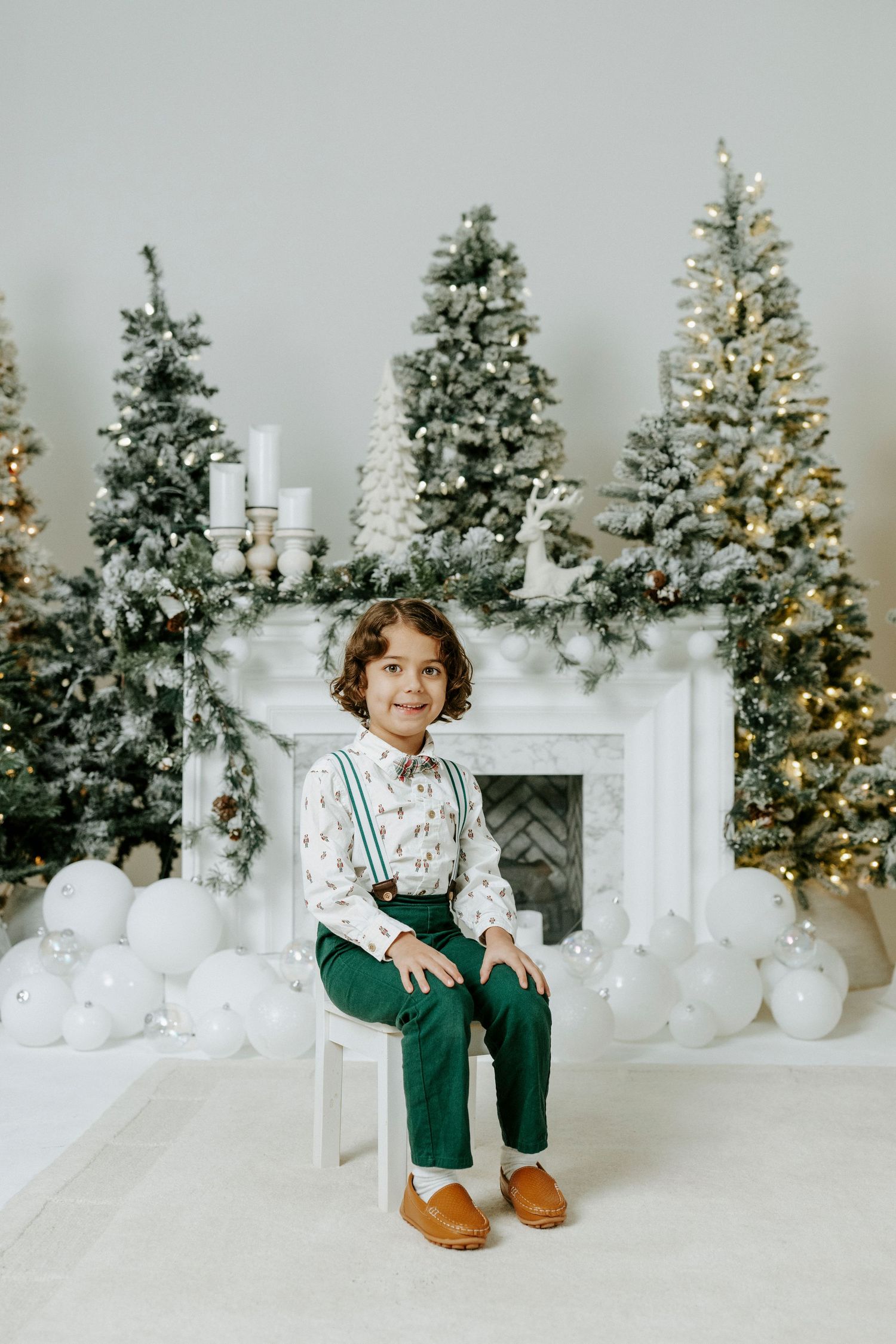 White Christmas studio setup with multiple decorated trees, a fireplace mantel, and festive holiday decor in soft lighting.
