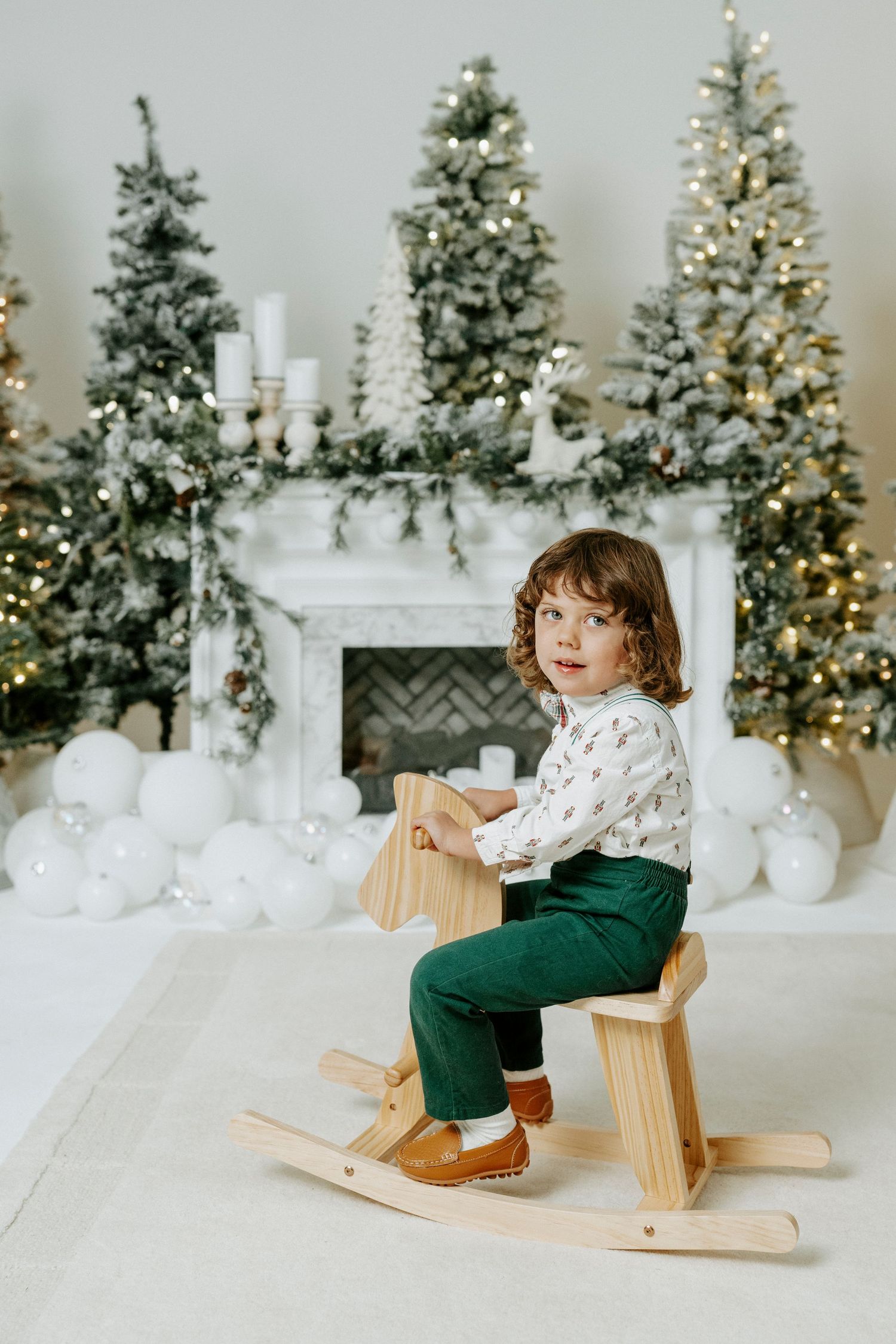 Child plays on wooden rocking horse in front of decorated Christmas trees and white winter decor setting.