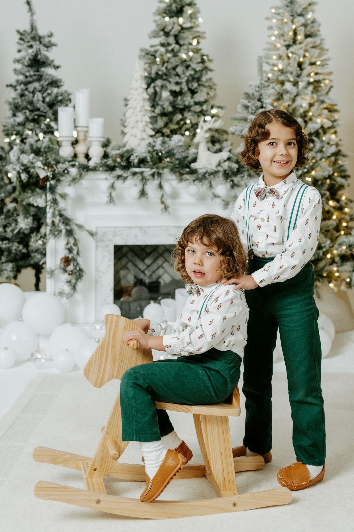 A child in a festive outfit plays on a wooden rocking horse in front of a decorated Christmas tree and fireplace.