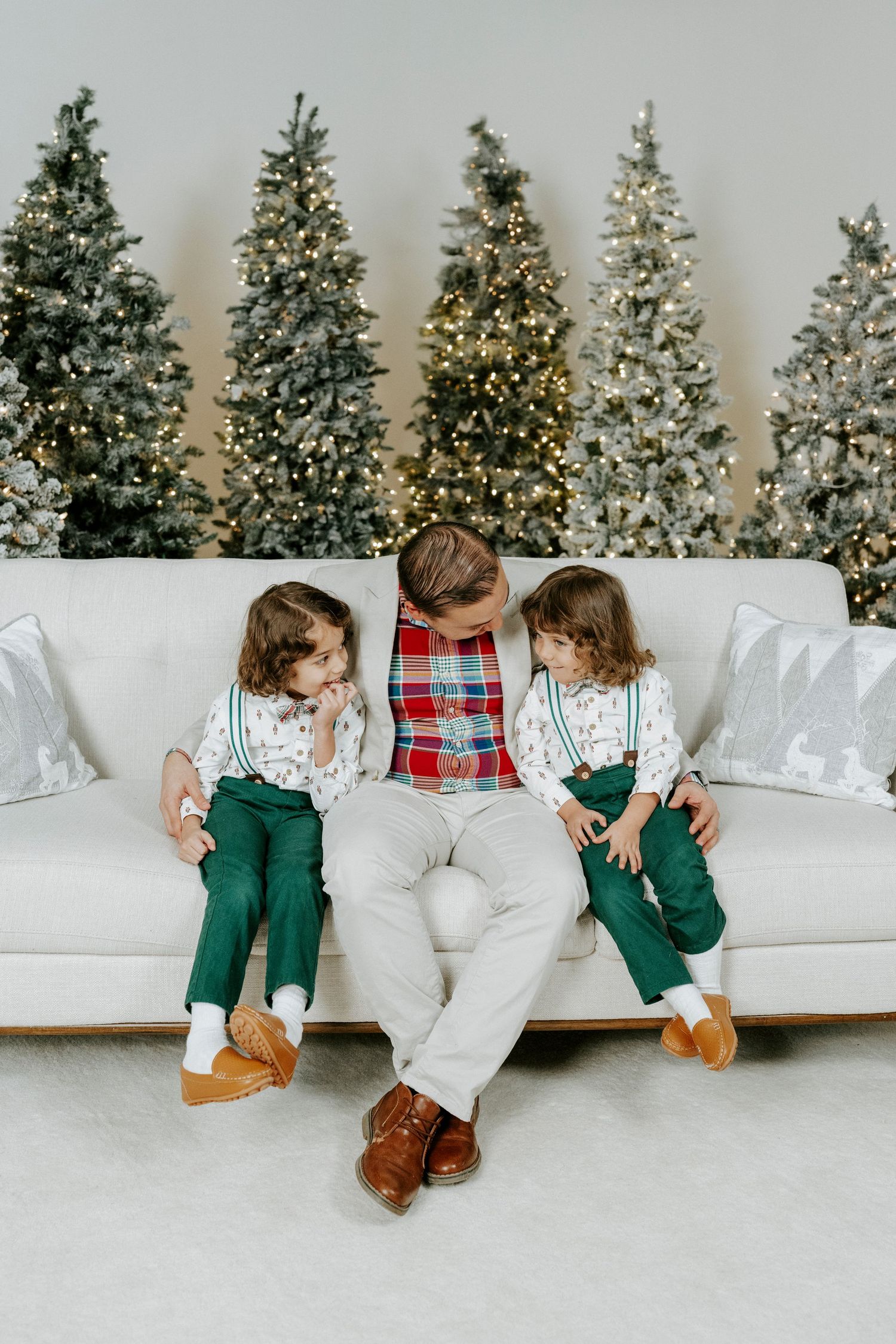 Children in matching white sweaters and green pants sit on white couch with Christmas trees in background.