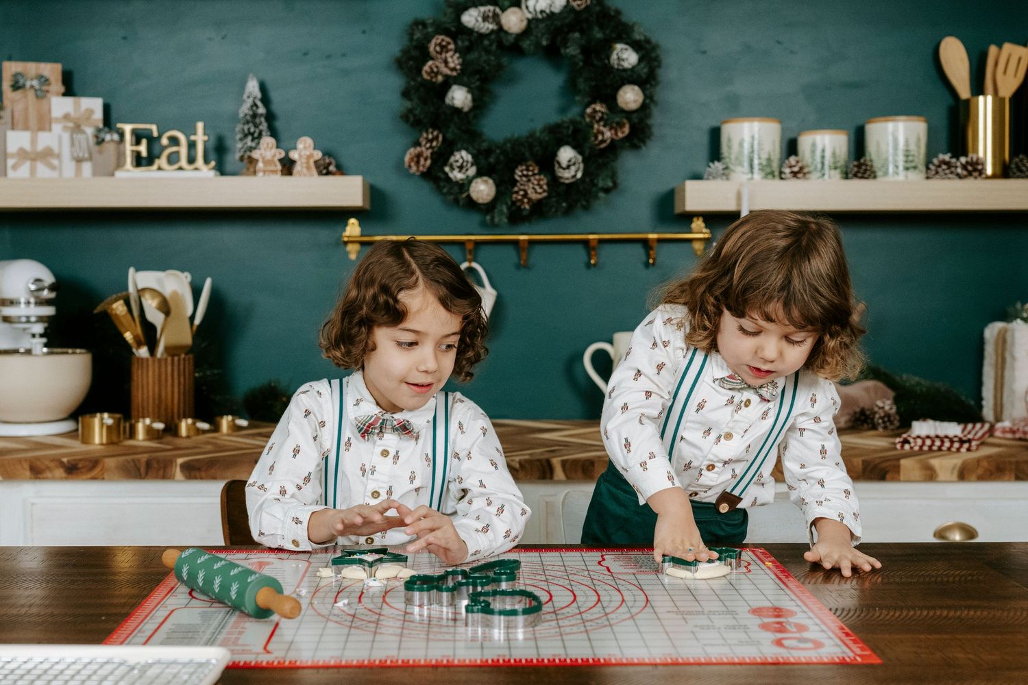 Children wearing floral aprons gather around a table to bake holiday cookies in a cozy kitchen with teal walls.