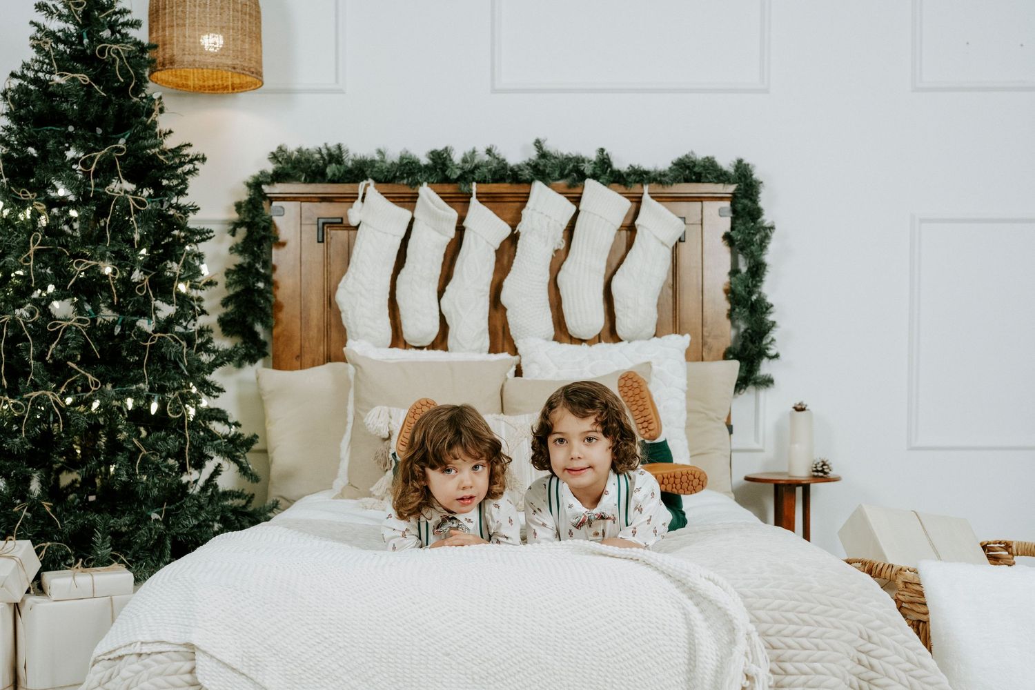 Two children sit together on a white bed with Christmas stockings hanging on the wall behind them.