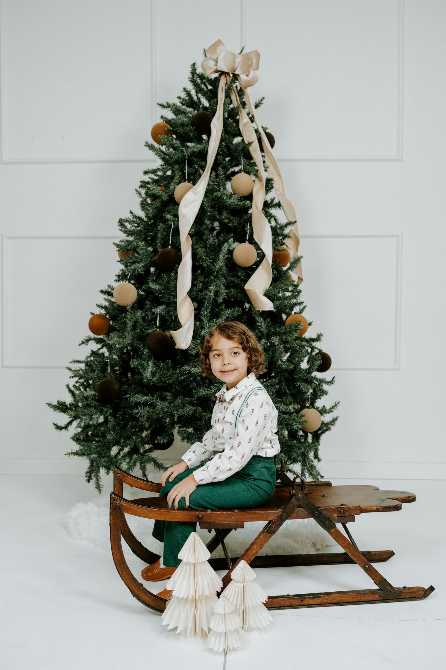 A decorated Christmas tree stands behind a vintage wooden sleigh in a minimalist white studio setting.
