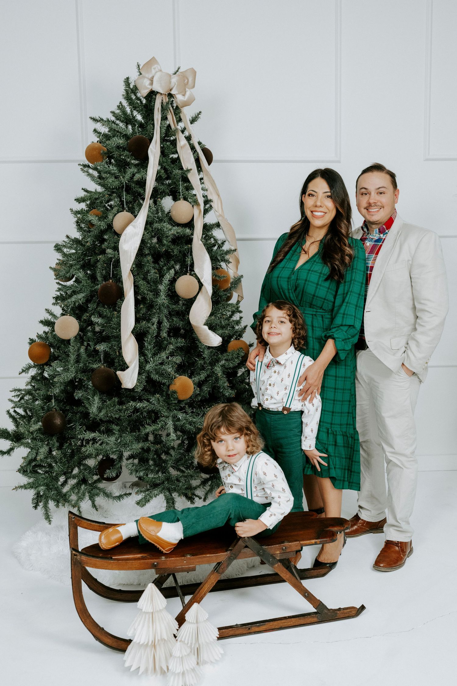 Family poses together in coordinated green and white outfits near a Christmas tree and wooden sleigh.