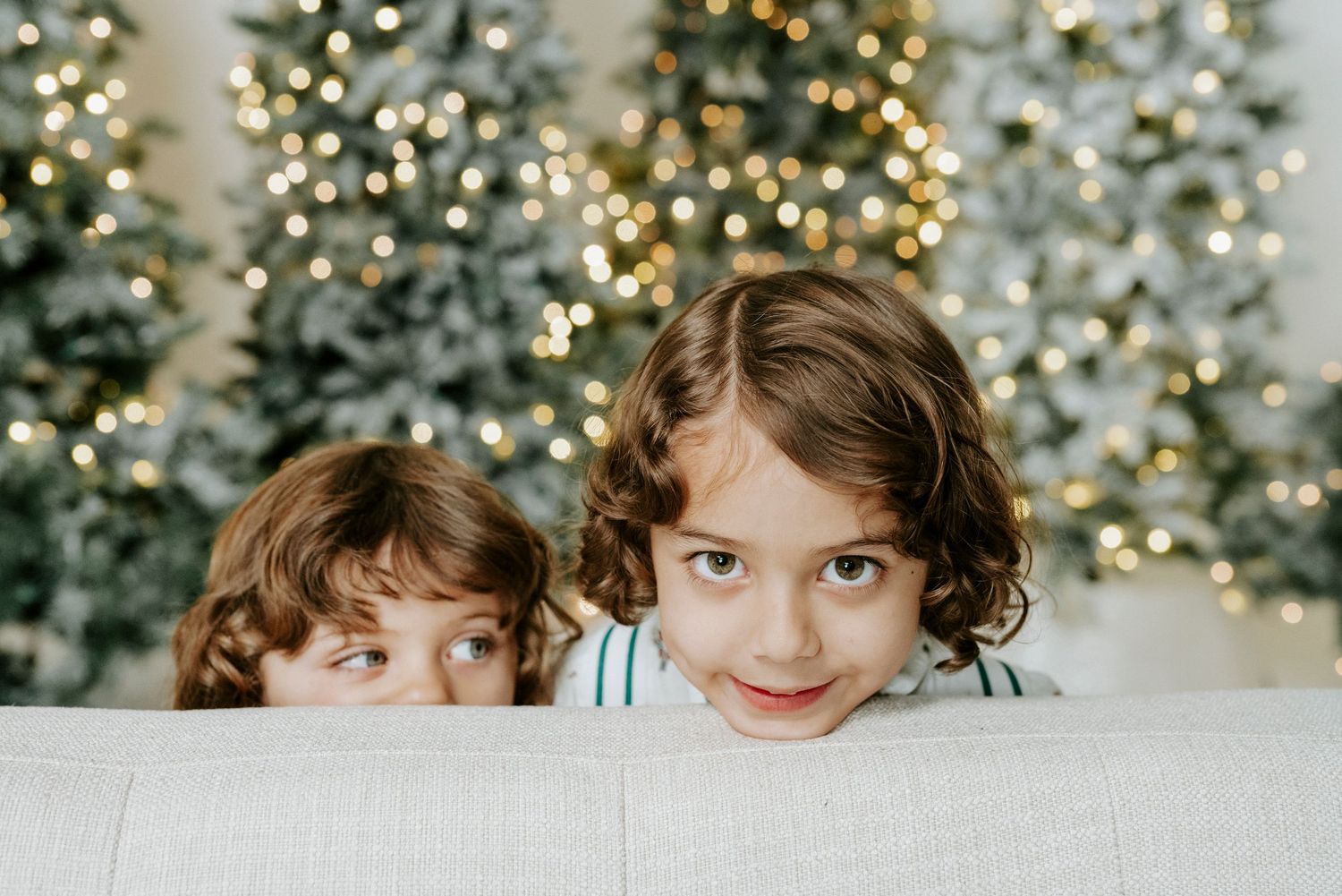 Two children peek playfully over a white couch with decorated Christmas trees and twinkling lights in the background.