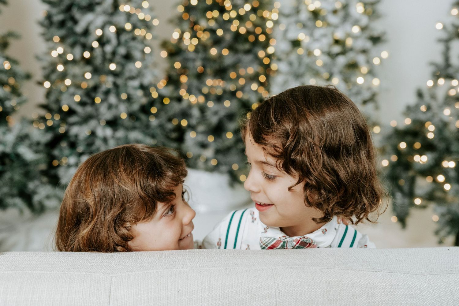Children share a sweet moment in front of a twinkling Christmas tree with bokeh lights in the background.