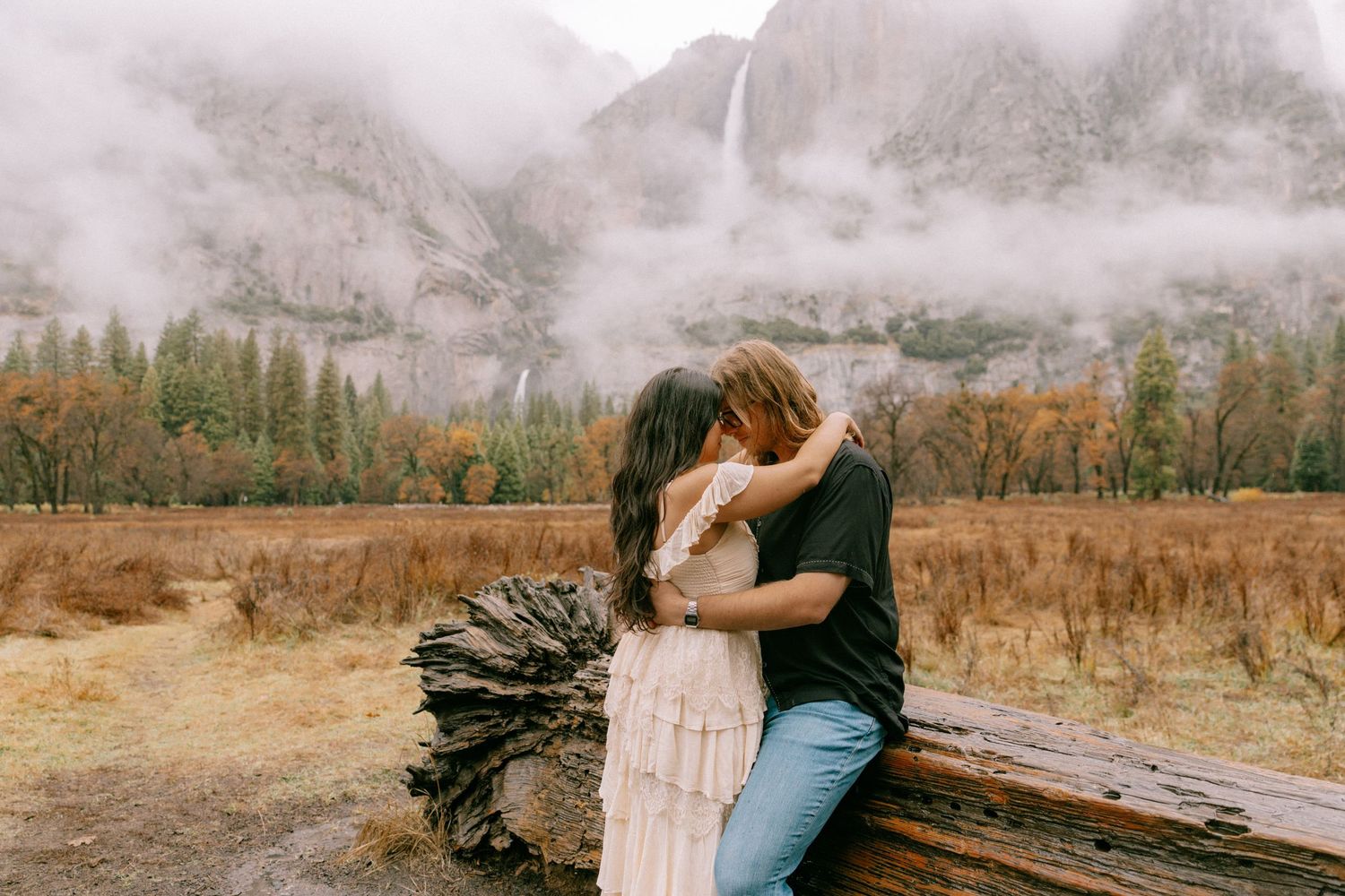 A couple embraces in a romantic moment against a misty autumn landscape with golden fields and trees in the background.