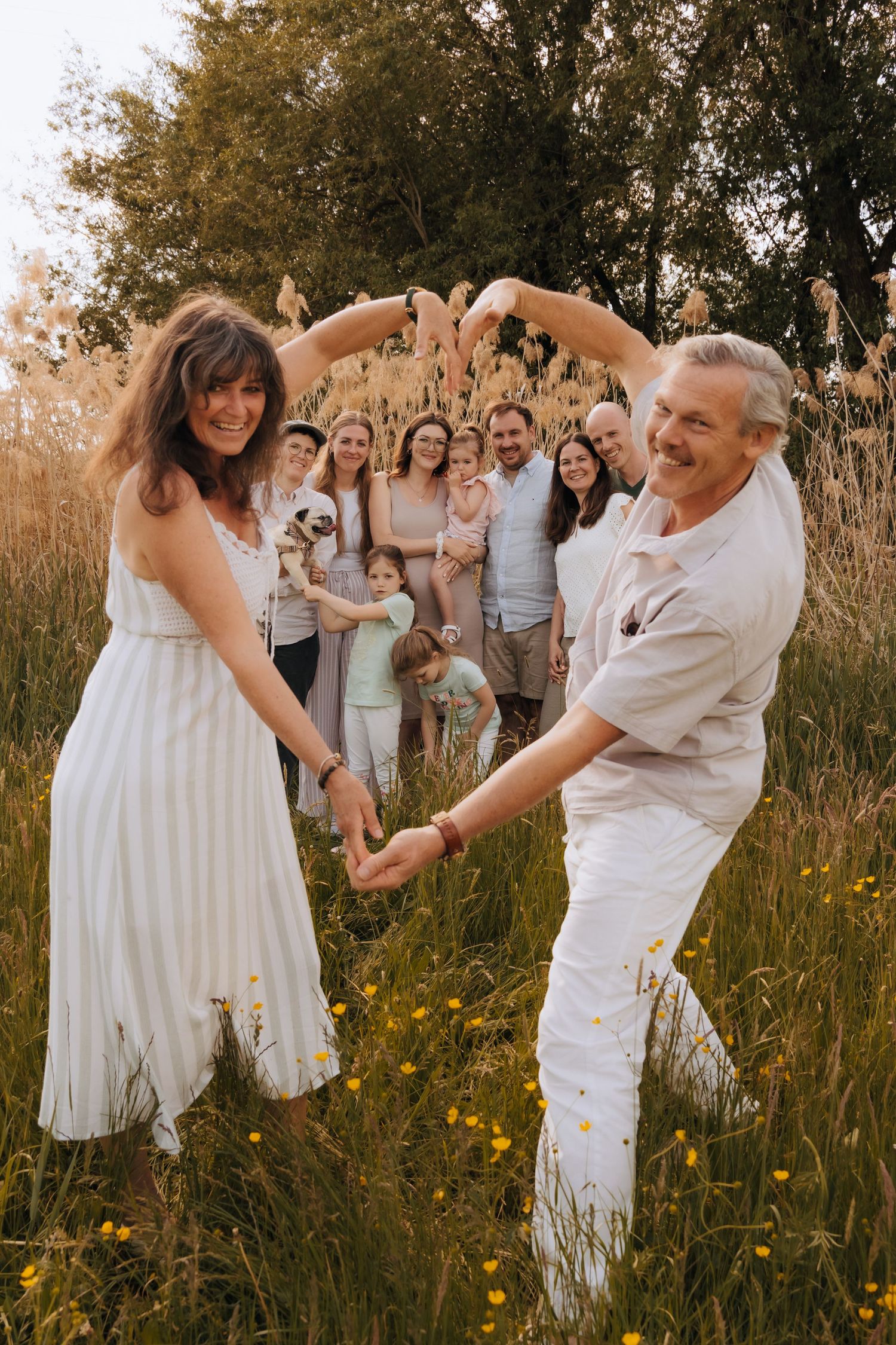 A couple in white outfits dancing in a golden field at sunset while wedding guests make heart shapes in the background.