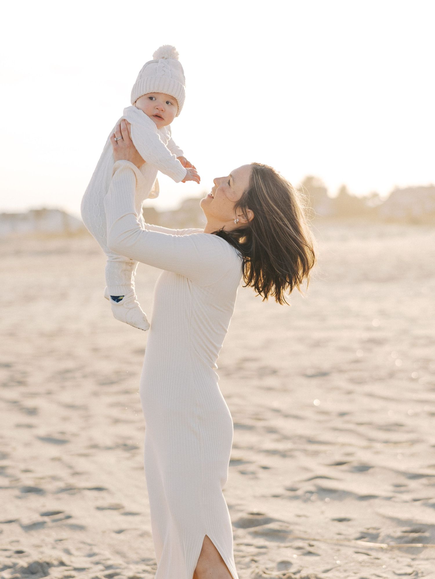 A young mother lifts her son into the air laughing together near the dunes during a fall beach photo session in New Jersey.