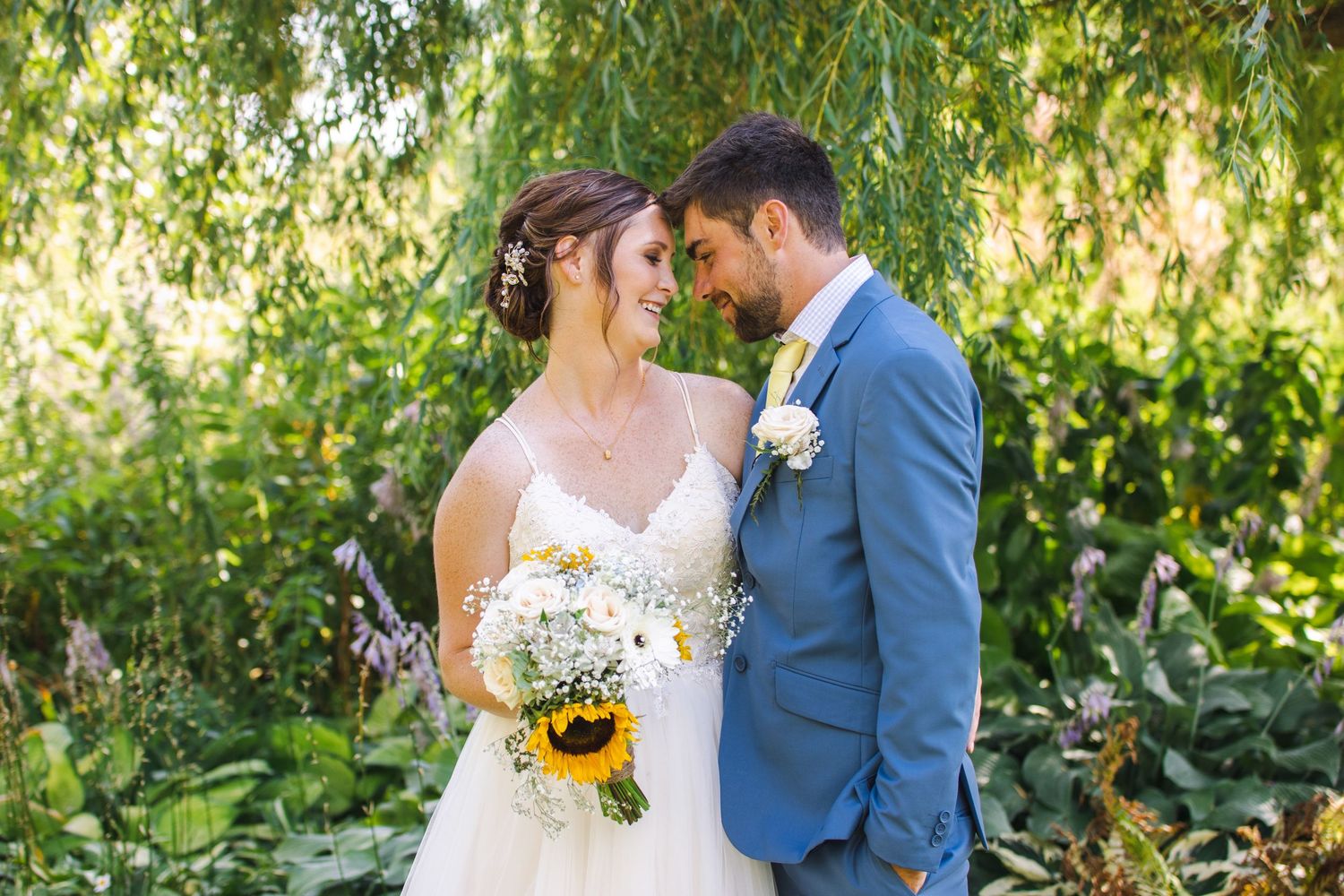 A romantic wedding portrait in a lush garden surrounded by greenery and sunlit foliage.