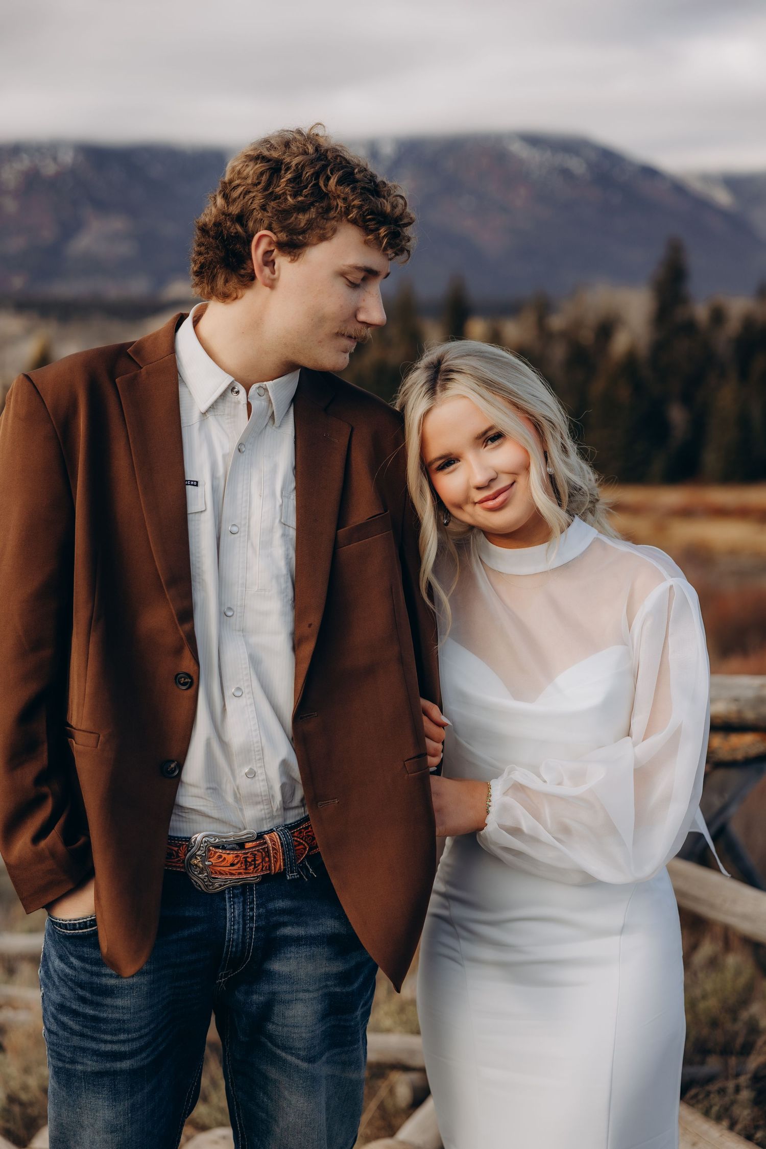 A couple in wedding attire poses together in a rustic mountain landscape with autumn colors in the background.