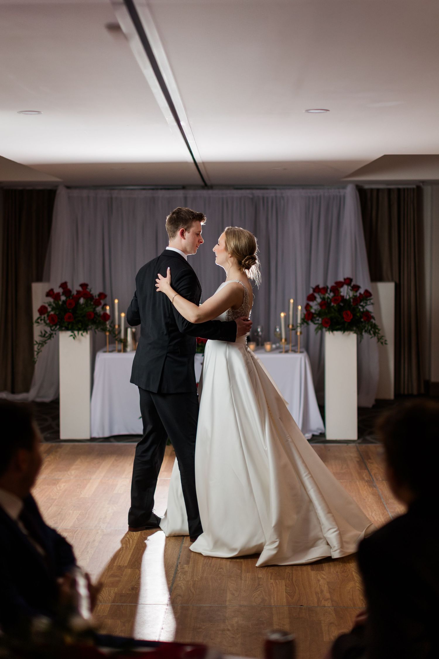 A couple shares their first dance as newlyweds in an elegant reception hall with red floral arrangements and white decor.