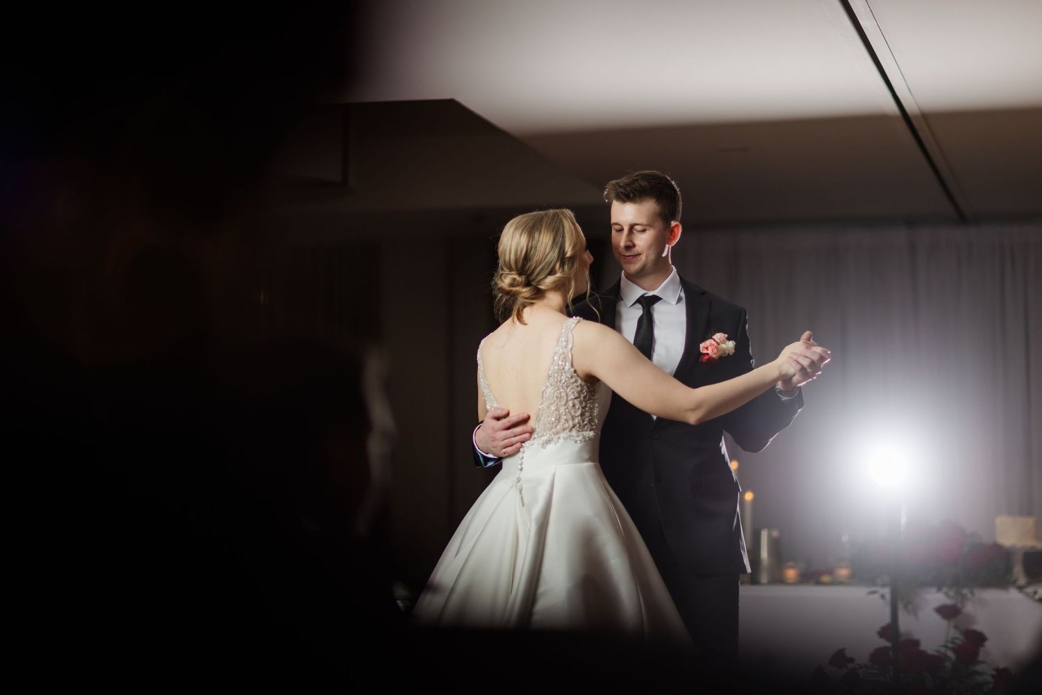 A couple dances together in a dimly lit reception venue with dramatic backlighting creating a silhouette effect.