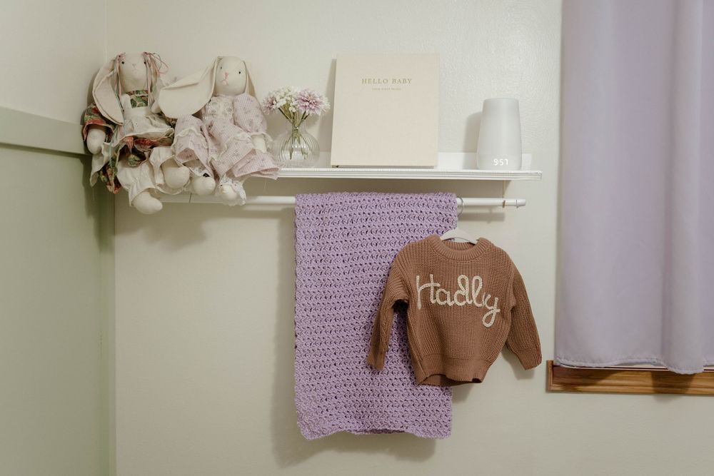 Cozy nursery corner with purple blanket and brown sweater hanging on white shelf.
