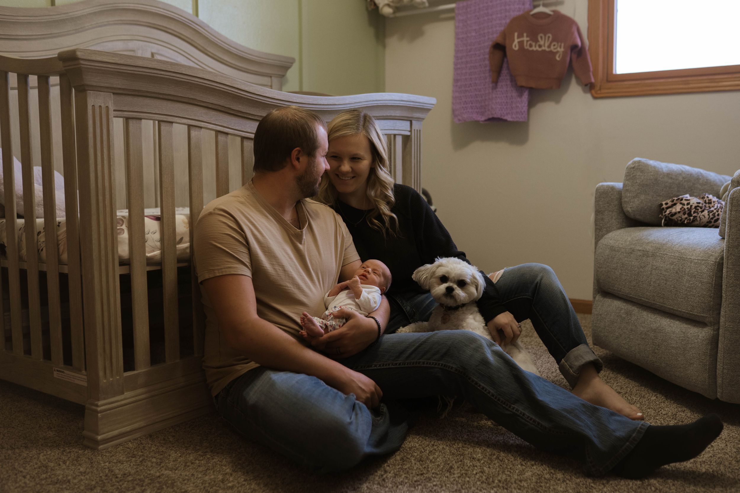 Family sitting on floor with white dog in nursery with wooden crib.